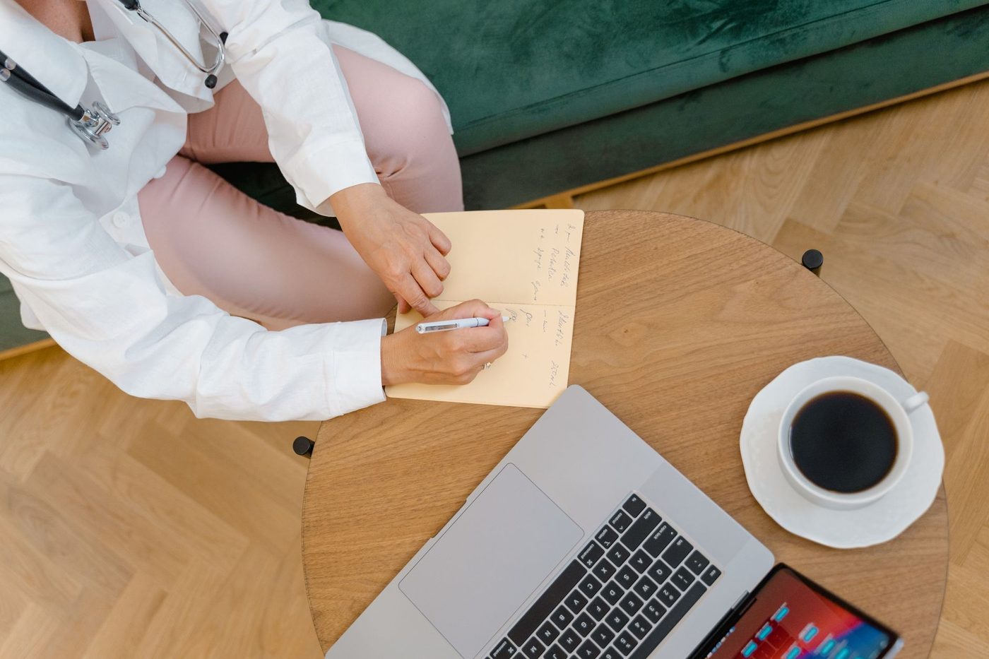 Overhead view of a clinician writing notes in a notebook at a table, with a laptop and a cup of coffee