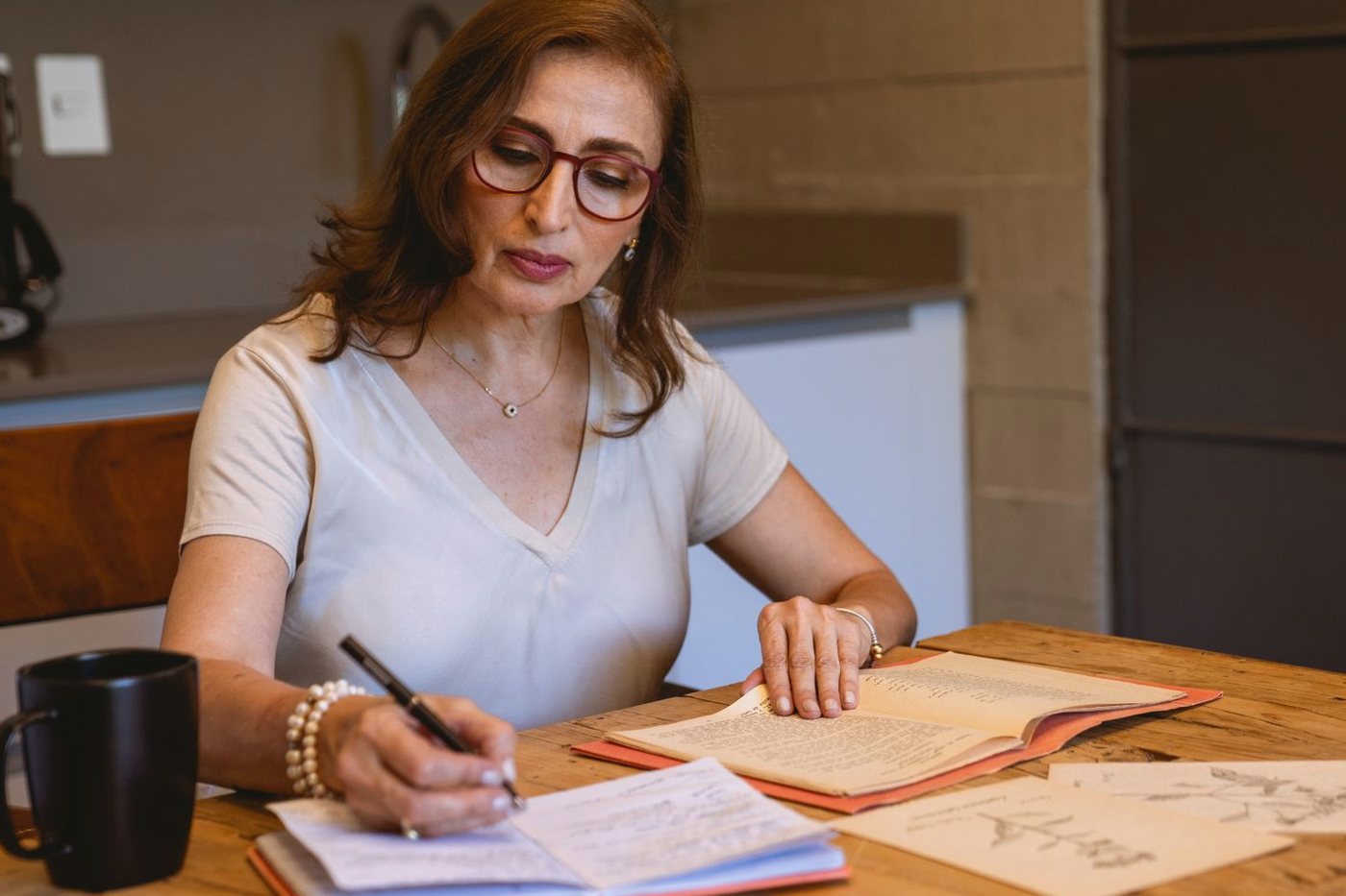 A woman writing notes in a notebook at a kitchen table, with papers and a cup of coffee